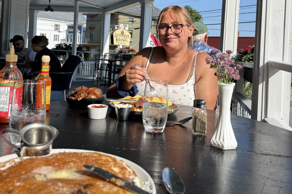 A woman sitting at an outdoor dining table enjoying a meal, with a plate of food in front of her, including pancakes and savory dishes. The table is set with condiments and a vase of flowers in a sunny setting.