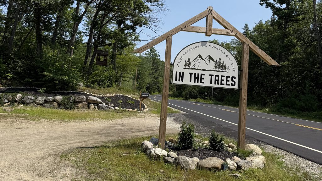 Wooden sign for 'In The Trees' featuring mountain graphics, surrounded by trees and landscaping at a roadside entrance.