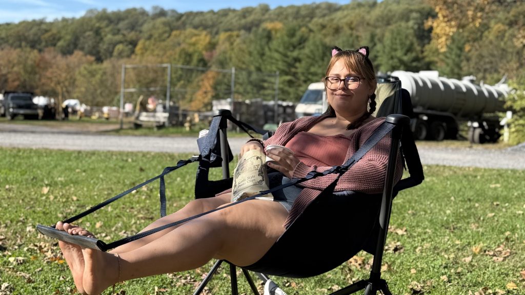 A person sitting in a camping chair outdoors, wearing a pink cardigan and cat ear headband, holding a cup, with green grass and trees in the background.