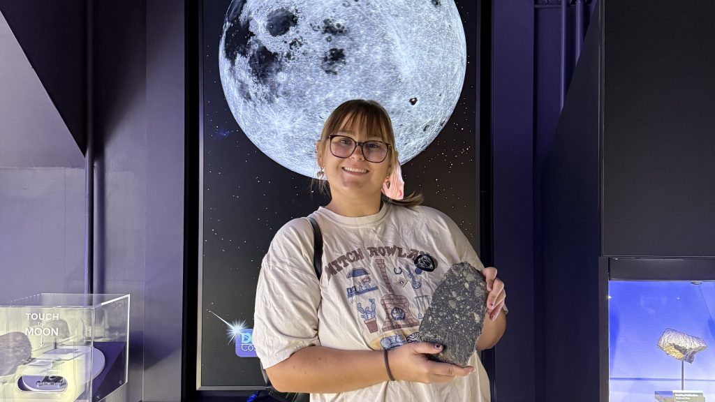 A person holding a rock, standing in front of a large moon backdrop in a museum exhibit.