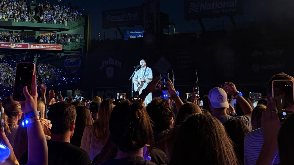 A musician performing on stage at a concert, with a large crowd holding up phones and wearing wristbands, illuminated by blue lights in a stadium setting.