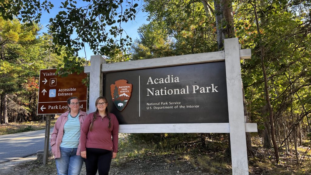 Two individuals stand next to the Acadia National Park sign, surrounded by trees. The sign indicates the park entrance and provides information about the National Park Service.